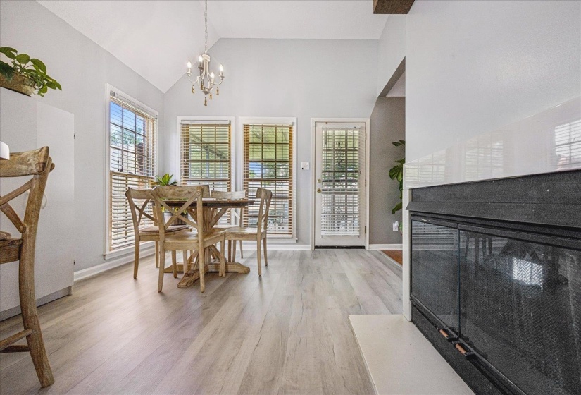 Vaulted ceiling with an elegant chandelier, three large windows, light wood-finish flooring, a glass-paneled door, and a built-in fireplace with a white hearth