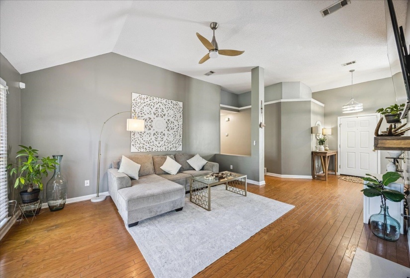 Spacious living area featuring a vaulted ceiling with a ceiling fan, wood-finish flooring, and a decorative front entry door with an overhead pendant light fixture
