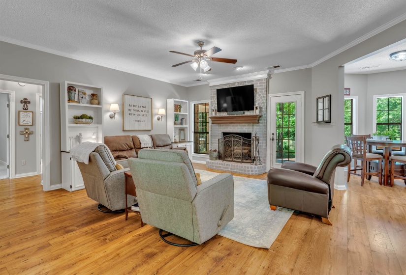 Spacious living area featuring light wood-finish flooring, a painted brick fireplace with a wood mantel, built-in shelving, an outdoor access door, and an open concept layout into an adjacent room