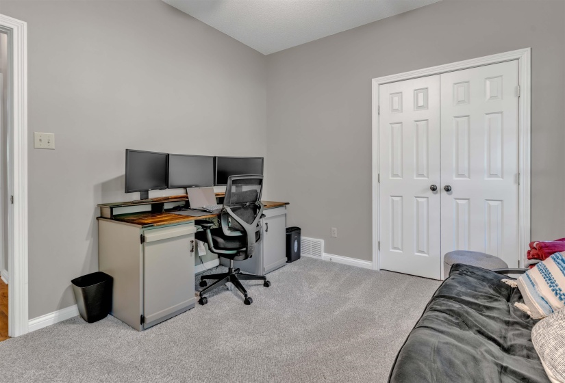 Dedicated room featuring a built-in wood-finish desk with storage cabinetry, light gray walls, plush gray carpeting, and white bi-fold closet doors