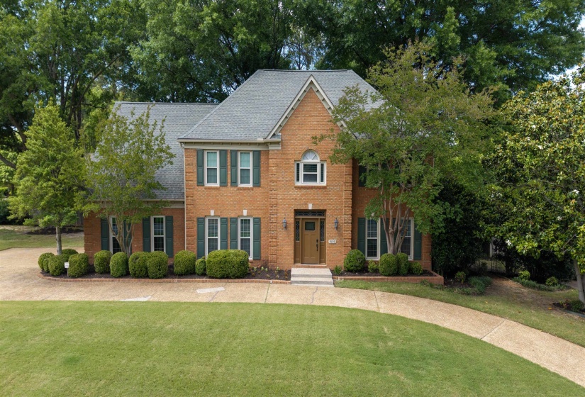 Brick exterior residence featuring a gabled roof, multiple windows with dark shutters, an arched entryway, and a paved circular driveway