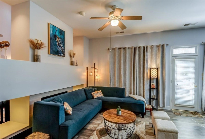 Open hearth area featuring a contemporary fireplace with a built-in shelf, wood-finish flooring, a ceiling fan, and a glass-paned door with blinds