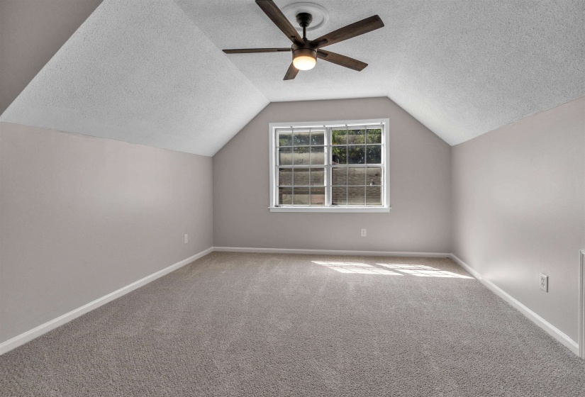 Vaulted ceiling room with an angled wall, featuring a multi-pane window, ceiling fan with light fixture, light gray wall paint, and gray carpet flooring