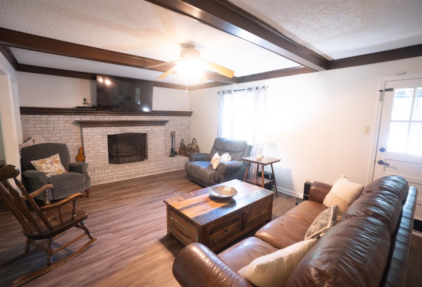 Living area featuring an exposed beam ceiling, white brick fireplace with dark wood mantel, wood-finish flooring, and a ceiling fan with light fixture