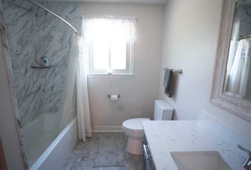 Full bathroom featuring a white tub with marble-patterned surround, a single window, a white toilet, a vanity with a white countertop and integrated sink, and gray tile flooring