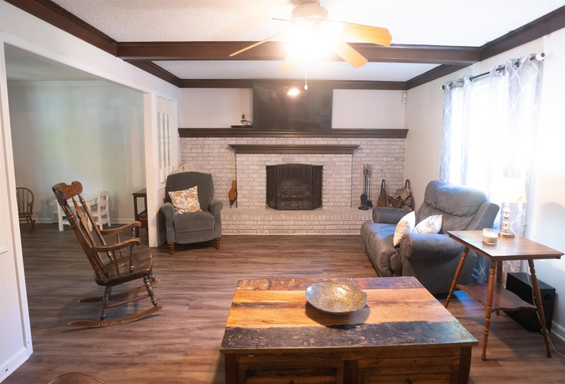 Spacious living area featuring wood-finish flooring, a white brick fireplace with a dark mantel, dark exposed ceiling beams, and a ceiling fan with integrated lighting