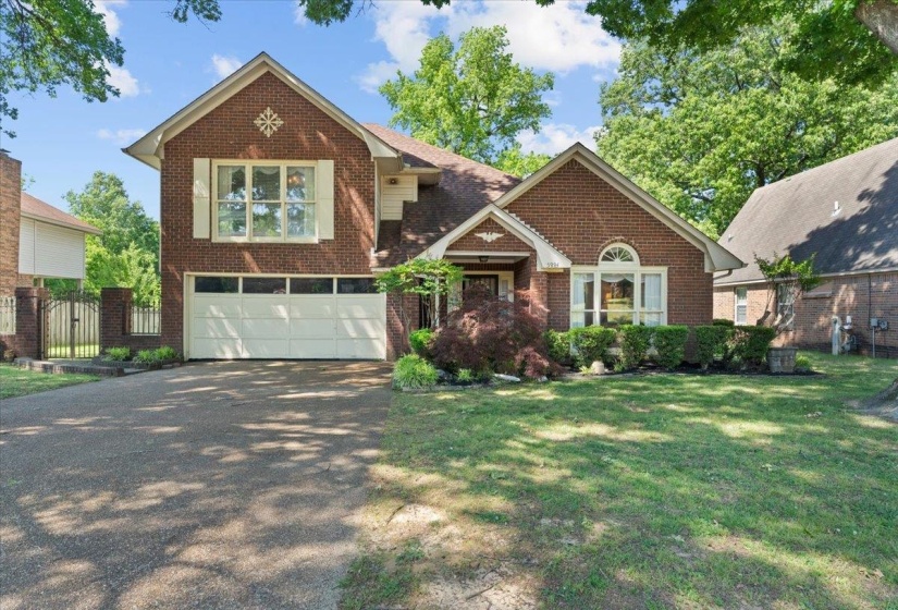 Traditional brick exterior featuring a two-car garage, established front landscaping, an arched window, decorative shutters, and mature trees