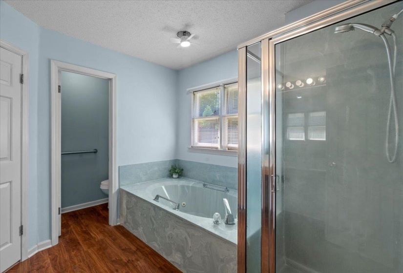 Bathroom featuring a corner soaking tub with a tiled surround, a glass-enclosed shower with a handheld showerhead, wood-finish flooring, a window with blinds, and light blue walls