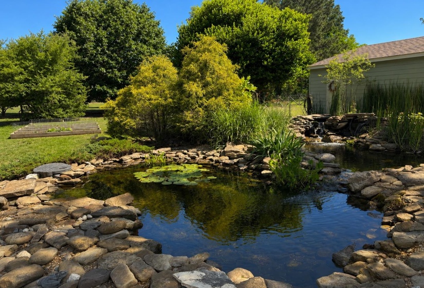Stone-lined pond with water feature, surrounded by lush greenery, mature trees, and a raised garden bed