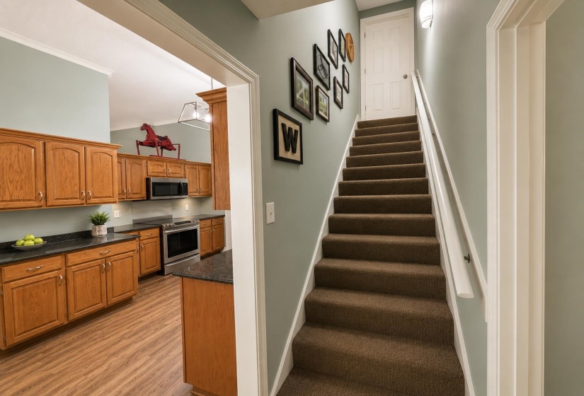 Carpeted staircase with white handrail and a doorway opening to a kitchen featuring wood-finish cabinetry, dark countertops, and stainless steel appliances