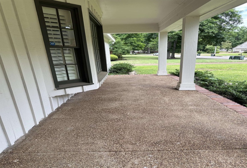 Front porch featuring an exposed aggregate concrete surface, white board and batten siding, dark framed windows with plantation shutters, brick edging, and white support columns