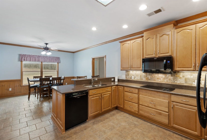 Spacious kitchen featuring light wood cabinetry, stone-finish countertops, black built-in appliances, and a tiled backsplash