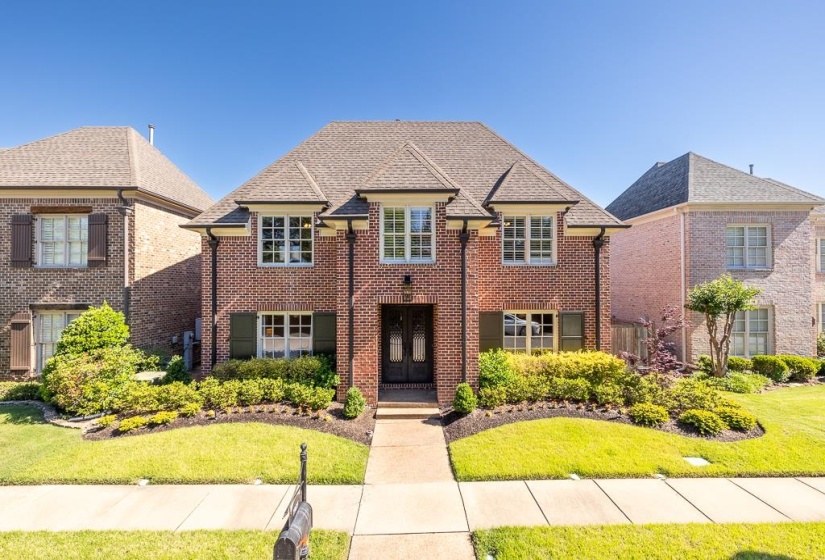Brick exterior featuring a gabled roofline, shuttered windows, a central entryway, a concrete walkway, and landscaped front yard with manicured lawn