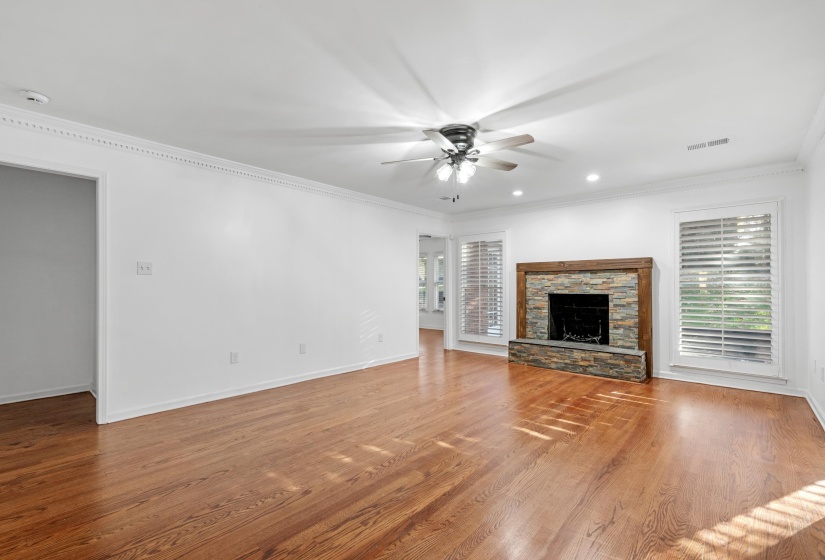 Expansive living area showcasing wood-finish flooring, a stone-clad fireplace with a wood mantel, and multiple windows with plantation shutters