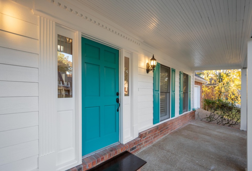 Covered front porch featuring a vibrant teal paneled door, white siding, brick foundation, and a wall-mounted exterior light fixture
