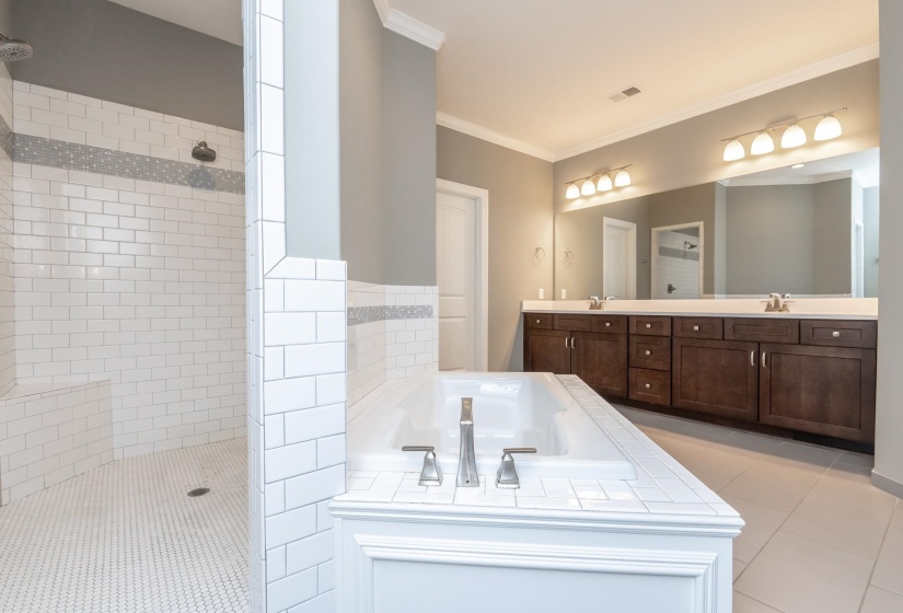 Spacious bathroom featuring a built-in soaking tub, a walk-in shower with subway tile and a mosaic accent, a double vanity with ample storage, a large mirror, and crown molding