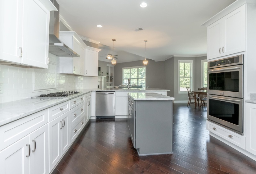Open-concept kitchen featuring dark wood-finish flooring, white shaker cabinetry, stainless steel appliances, and a central gray island with a light-toned countertop