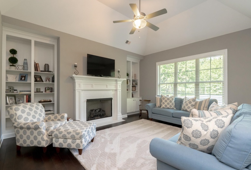 Living area featuring a vaulted ceiling with a ceiling fan, a fireplace with a white mantel, built-in shelving, wood-finish flooring, and a large window with blinds