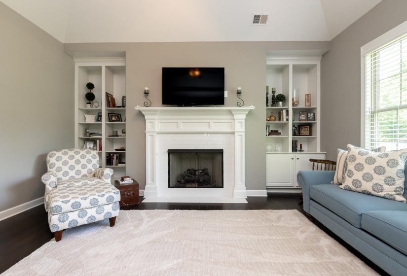 Vaulted ceiling living area featuring a prominent white mantel fireplace, dark wood-finish flooring, and built-in shelving with lower cabinetry