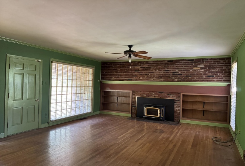 Living area featuring wood-finish flooring, a brick accent wall, built-in shelving, a wood-burning stove, and a ceiling fan