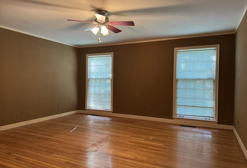 Spacious room featuring hardwood flooring, two large windows with blinds, a ceiling fan with integrated lighting, crown molding, and baseboard trim
