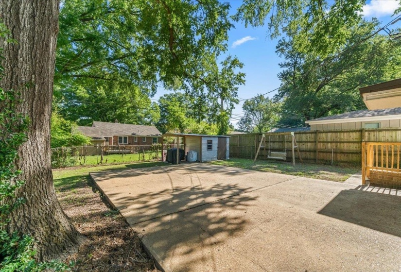 Expansive concrete patio with a wooden privacy fence, storage shed, and mature trees providing natural shade