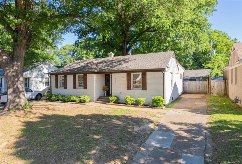 Exterior features a white painted brick facade with brown decorative shutters, a gabled roof, and a mature tree providing shade to the front yard
