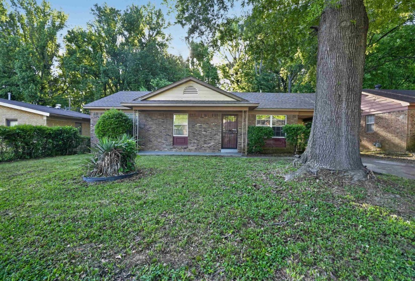 Brick facade ranch-style home featuring a gabled roofline, mature trees, and established shrubbery