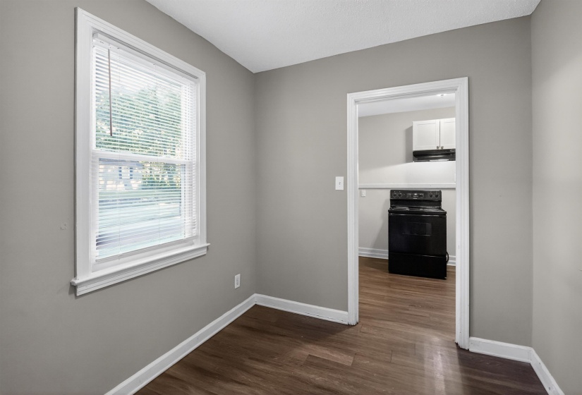 Windowed room featuring wood-finish flooring, light grey painted walls, white trim, and a doorway to a kitchen with a black electric range and white upper cabinetry