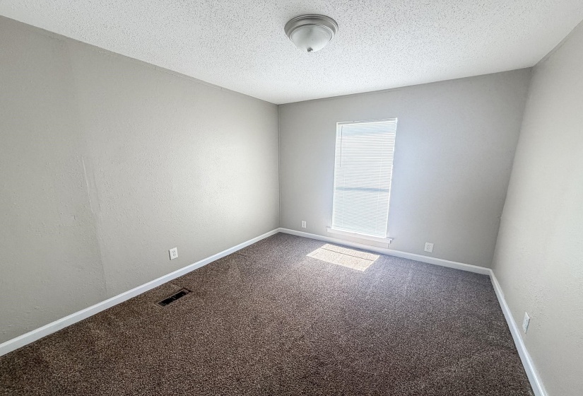 Neutral tone wall paint, textured ceiling, brown patterned carpet, white baseboards, and a single window with blinds