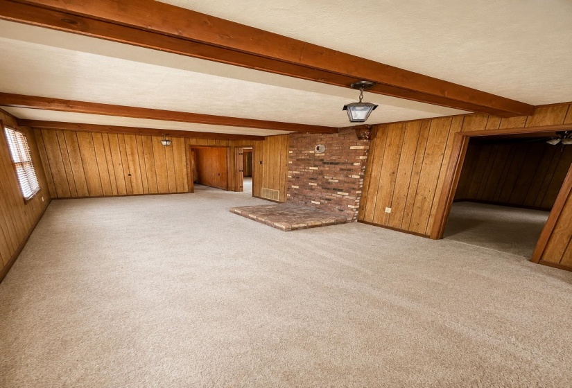 Room featuring beige carpet flooring, wood-paneled walls, exposed wood ceiling beams, and a brick fireplace