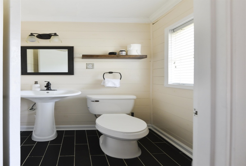 Compact bathroom featuring black plank flooring, light-toned shiplap walls, a pedestal sink with a matte black faucet, and a wall-mounted wood-finish shelf