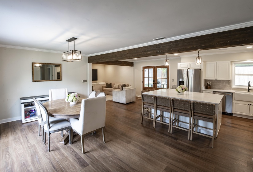 Open-concept living space featuring wood-finish flooring, exposed ceiling beams, a central kitchen island with seating, white cabinetry, and stainless steel appliances