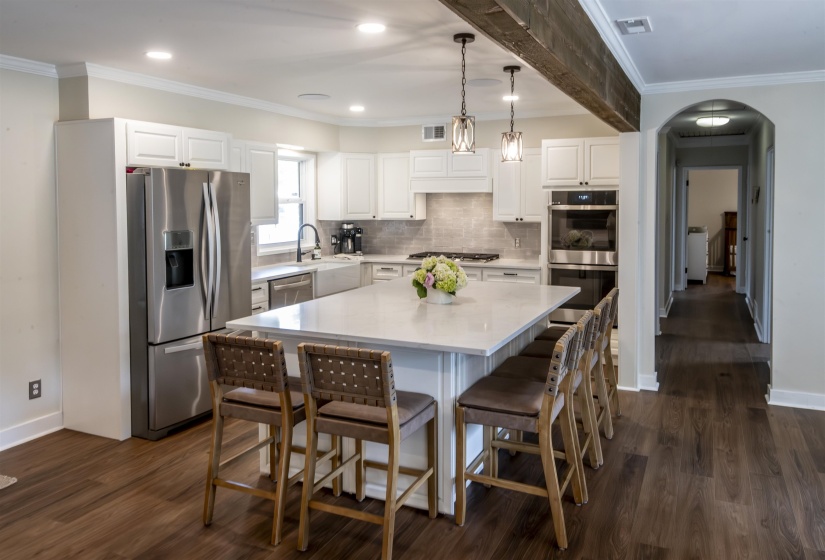 Open-concept kitchen featuring an expansive central island with seating, white shaker cabinetry, stainless steel appliances, and wood-finish flooring