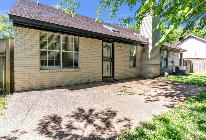 Concrete patio with a light brick exterior, dark-framed windows with blinds, a security screen door, and a brick chimney