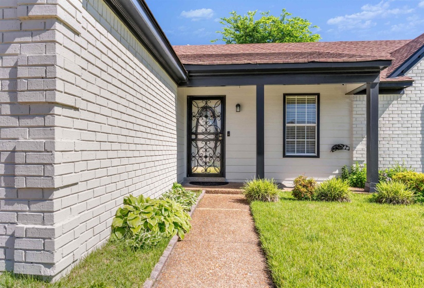Front entry featuring a security screen door with ornate ironwork, a covered porch with dark columns, a single-hung window with white blinds, white siding, and light-colored brick
