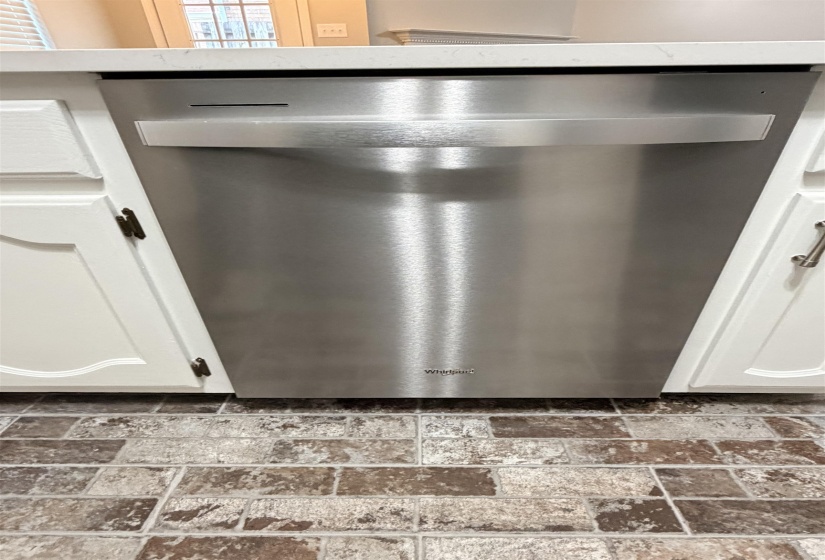 Stainless steel dishwasher integrated into white cabinetry with a light-colored countertop and multi-toned tile flooring