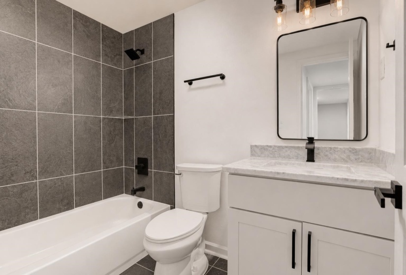 Bathroom featuring a white bathtub with dark gray tile surround, matte black shower fixtures, a white vanity with a light-colored countertop, a rectangular mirror, and a three-light vanity fixture
