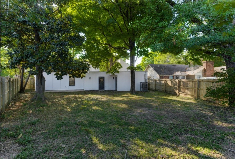 Spacious rear yard featuring extensive grassy areas, mature shade trees, a full privacy fence, and a rear door entry point