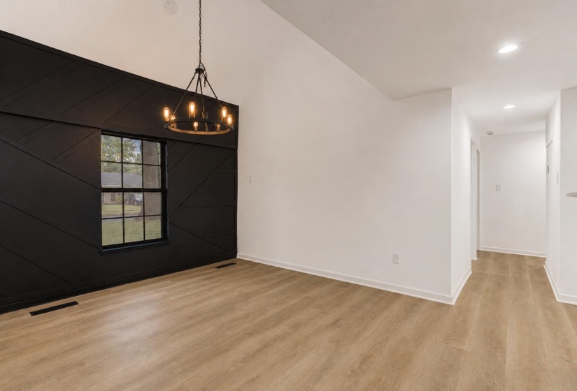 Vaulted ceiling with an exposed beam-style chandelier, contrasting accent wall with geometric paneling, double-hung window, wood-finish flooring, and recessed lighting