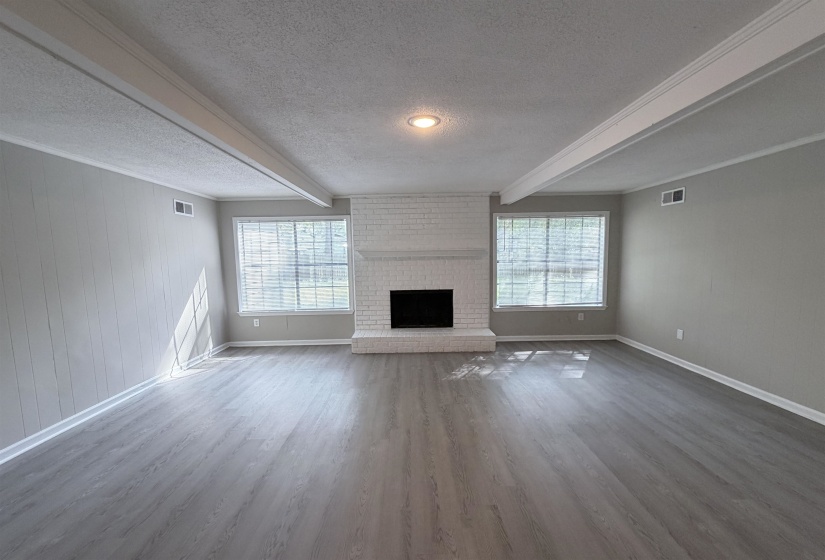Spacious interior featuring wood-finish flooring, a painted brick fireplace with hearth, two large windows, white trim, and exposed ceiling beams