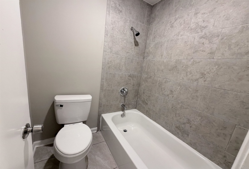 Full tile surround bathtub and shower combination with a bright white toilet, light gray painted walls, and coordinated gray tile flooring