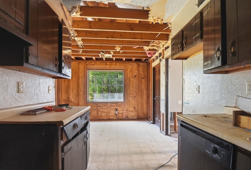Kitchen space featuring exposed ceiling joists, wood paneling, a large window, dark upper and lower cabinetry, and a built-in black dishwasher