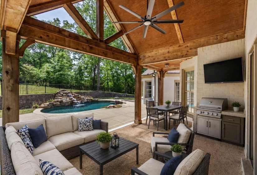 Covered Patio with vaulted wood ceiling, exposed beams, and ceiling fan, enhanced by outdoor living and dining areas with built-in grill—overlooking pool with waterfall feature for effortless entertaining. Virtually staged photo.