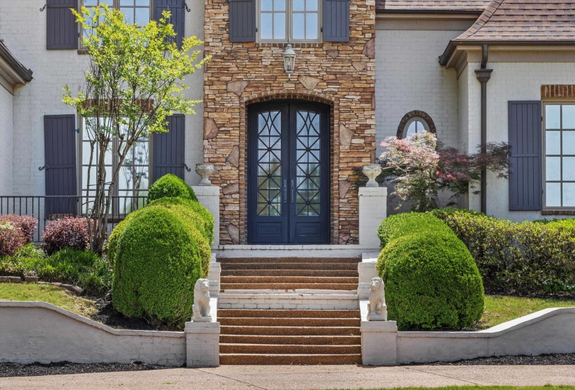Stunning curb appeal with custom iron double doors framed by stone, manicured landscaping, classic shutters, and elegant entry steps. Warm and welcoming impression.