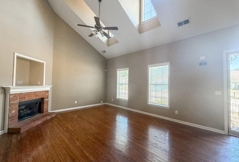 Spacious living area featuring high vaulted ceilings with skylights, rich wood-finish flooring, and a brick fireplace with a white mantel