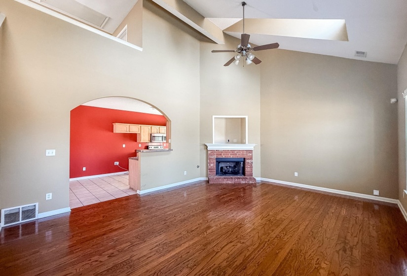 Spacious living area featuring high vaulted ceilings, wood-finish flooring, a brick fireplace, and an arched doorway leading to a kitchen with light wood cabinetry