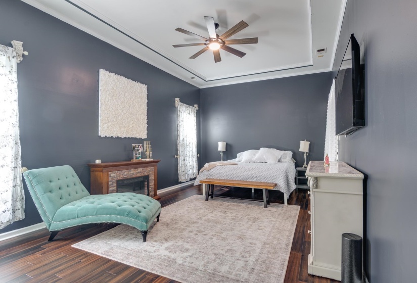 Recessed tray ceiling with crown molding, dark wood-finish flooring, and a stone-clad fireplace with a wood mantle