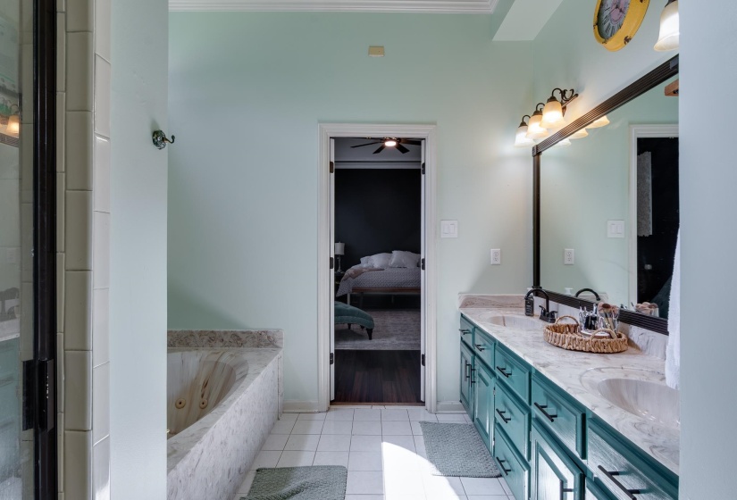 Bathroom featuring a dual vanity with a light-colored countertop and teal cabinetry, a large mirror with integrated lighting, a built-in tub with stone-look surround, and white tile flooring