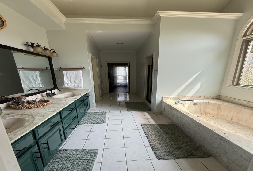 Spacious bathroom featuring a dual vanity with green cabinetry and light-toned countertops, a large soaking tub with a stone-look surround, a tiled floor, and crown molding details